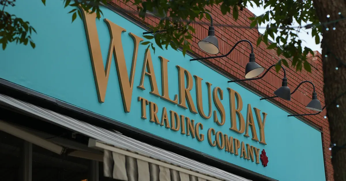 The Walrus Bay Trading Company storefront: gold lettering on a teal sign, lit by gooseneck lamps, under tree branches.